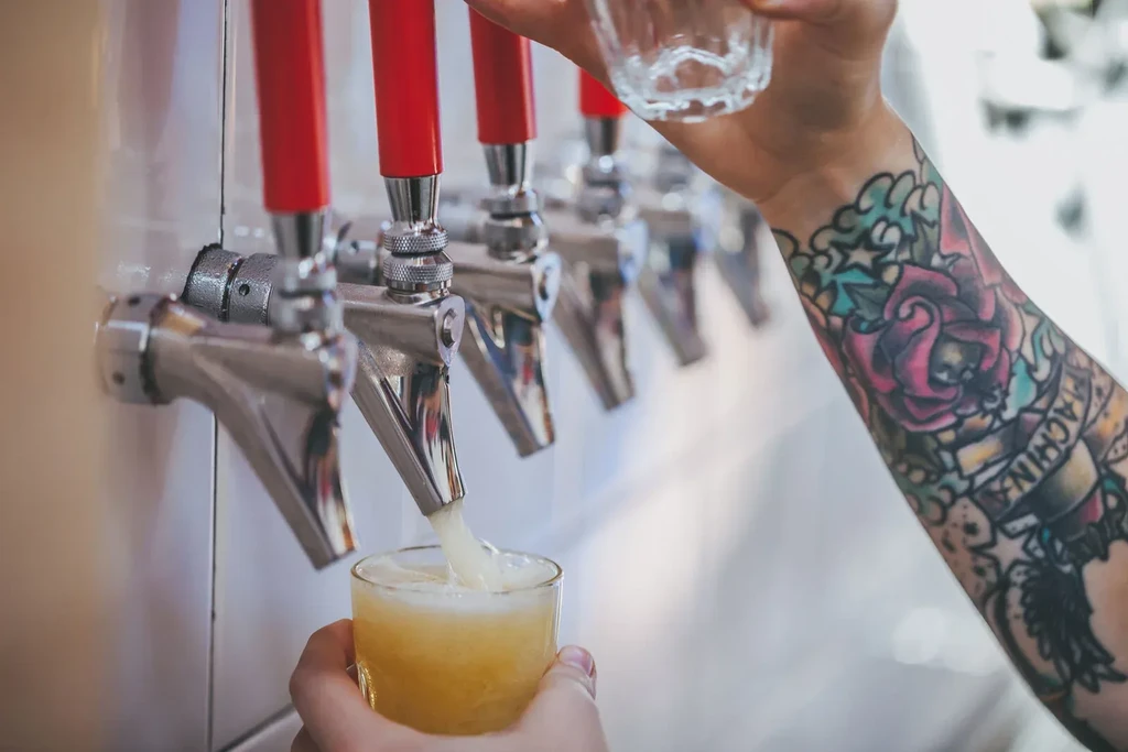 Tattooed hand pours frothy beer from a tap into a glass.