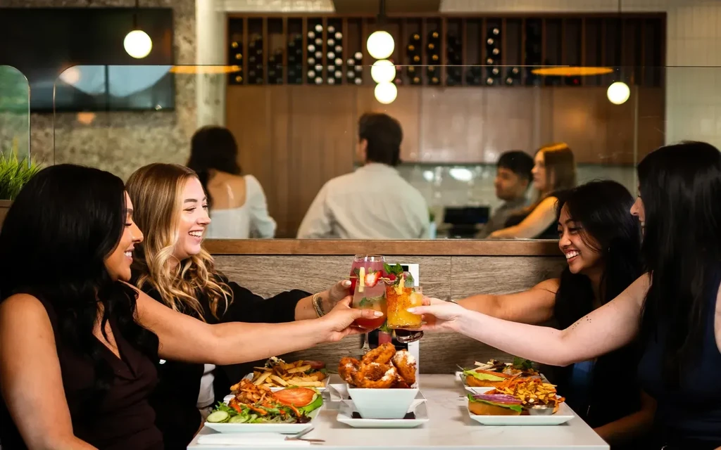Four young women clink cocktails and smile over food in a restaurant.