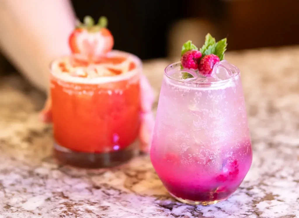 Sparkling pink and red drinks with garnishes on a stone counter.