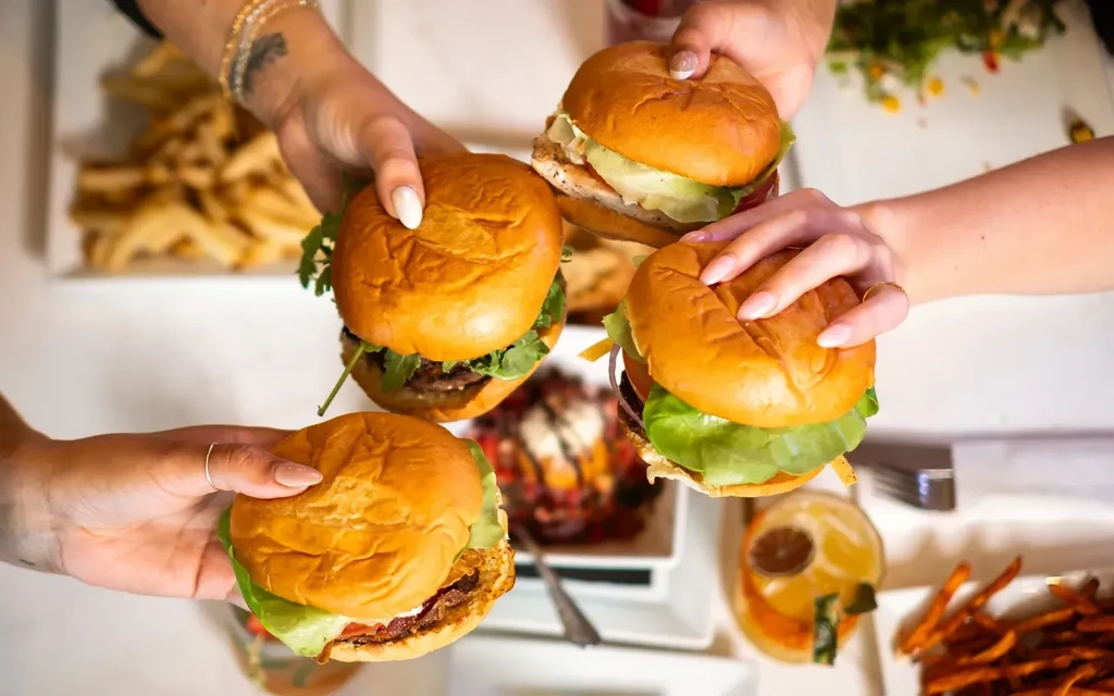 Multiple hands holding four different burgers and sandwiches in a restaurant.