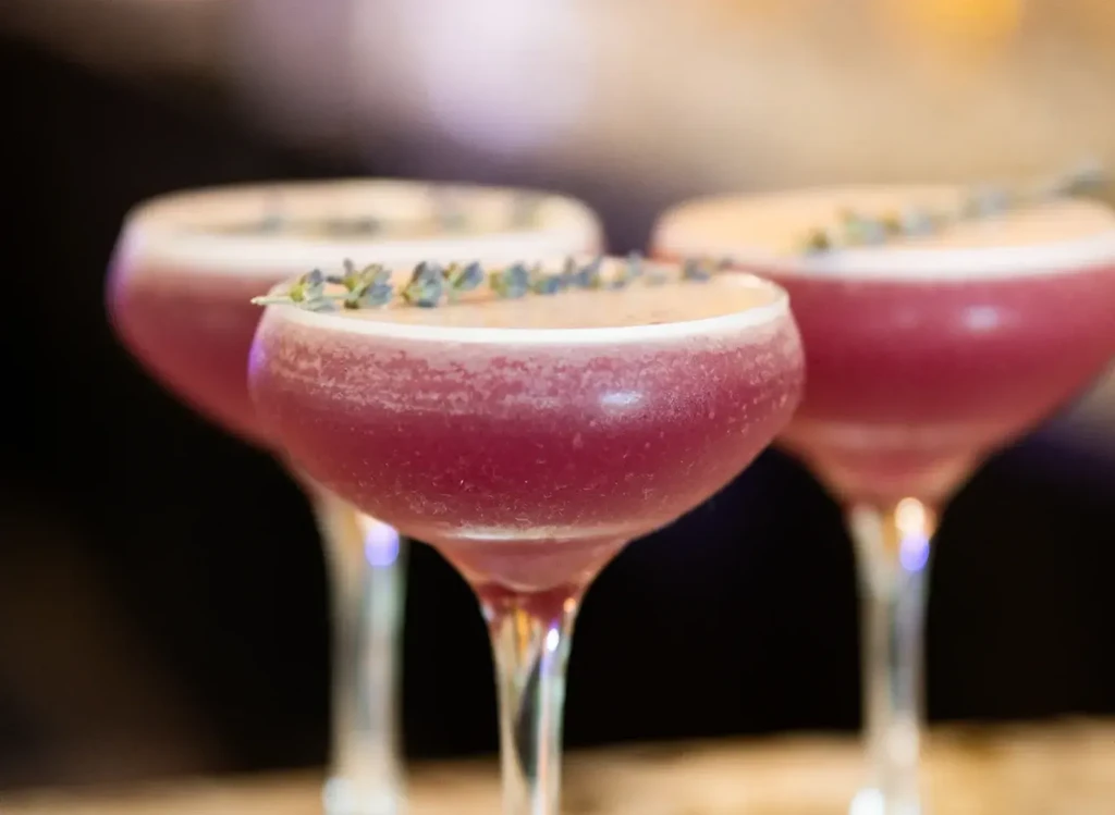 Fuchsia cocktail in coupe glass, foam, lavender, two similar drinks.