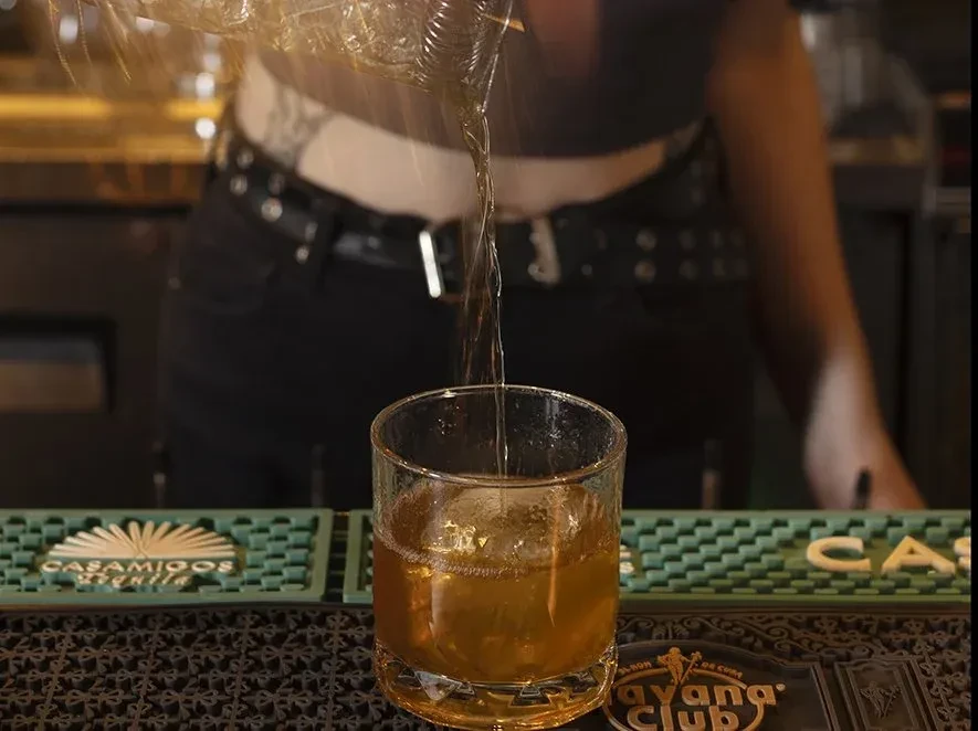 Bartender pours amber drink into a rocks glass with spherical ice.