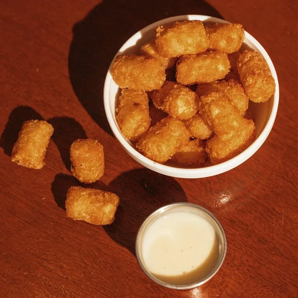 Bowl of golden tater tots, some spilled, with dipping sauce.