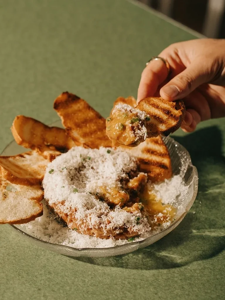 Rustic dip topped with cheese, served with grilled bread and a hand eating.