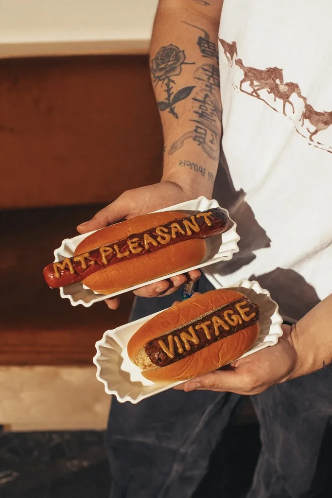 Hot dogs labeled 'MT PLEASANT' and 'VINTAGE' held by a tattooed arm.