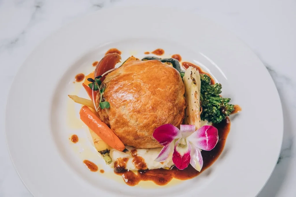 Golden-brown flaky pastry, vegetables, sauce, and pink orchid on a plate.