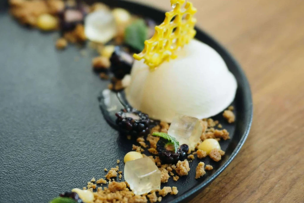 Plated white dome dessert with yellow honeycomb tuile, berries, and crumbles.