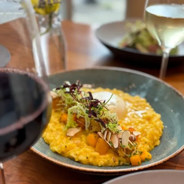 Yellow risotto in teal bowl with garnishes, wine on wooden table.