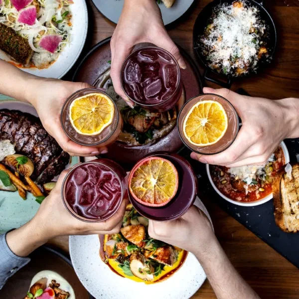 Five hands toast garnished drinks over a festive, food-filled table.