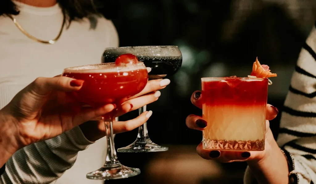 Three hands hold three colorful, garnished cocktails in dim lighting.