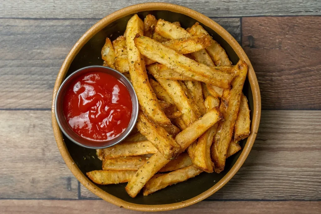 Overhead: Seasoned skin-on fries and ketchup in a bowl on wood.