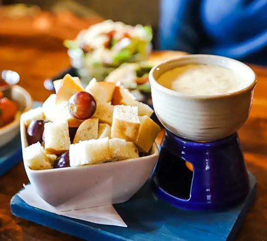 Cheese fondue, bread cubes, and red grapes on a blue stand.
