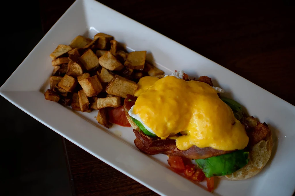 Breakfast plate with Eggs Benedict, bacon, avocado, and home fries.