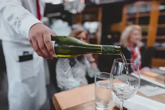 Server's hand pouring white wine from a green bottle into a glass.