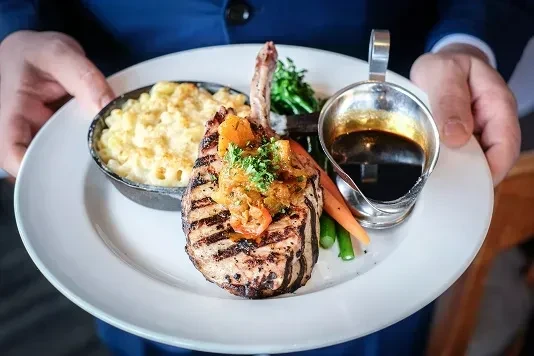 Server holding grilled bone-in pork chop, mac and cheese, vegetables, dark sauce.