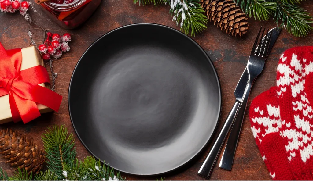 Christmas table setting with black plate, pine, berries, wine, gift, and mitten.