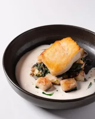 Seared white fish, scallops, vegetables, and creamy sauce in a dark bowl.