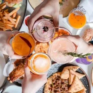 Hands clinking colorful drinks and beverages at a table with food.