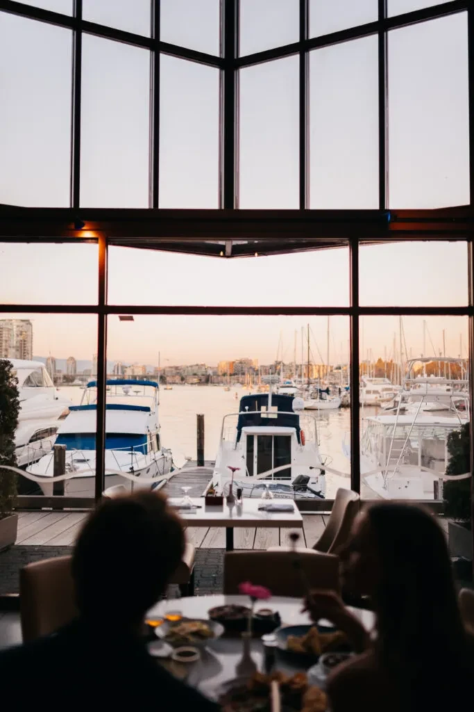 Silhouetted diners overlook a golden hour marina and city skyline.