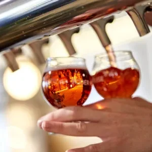 Hands filling two small glasses with amber beer from taps.