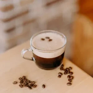Clear glass mug of coffee with foam, three beans, and scattered beans.