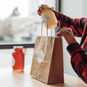 Plaid-shirted hands placing a yellow jar into a brown takeout bag.