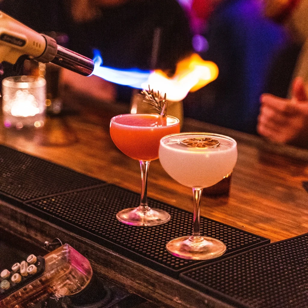 Two cocktails in coupe glasses being flamed on a dim bar.
