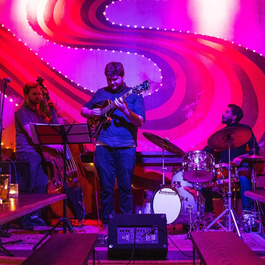 Trio of male musicians plays guitar, bass, drums under magenta lights.
