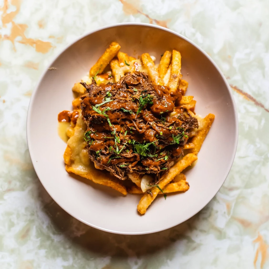 Dish of fries smothered in cheese, dark shredded meat, and herbs.