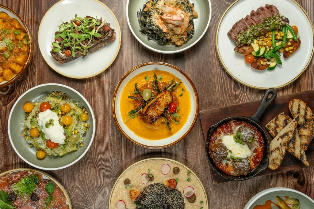 Overhead flat lay of ten diverse gourmet dishes on a dark table.