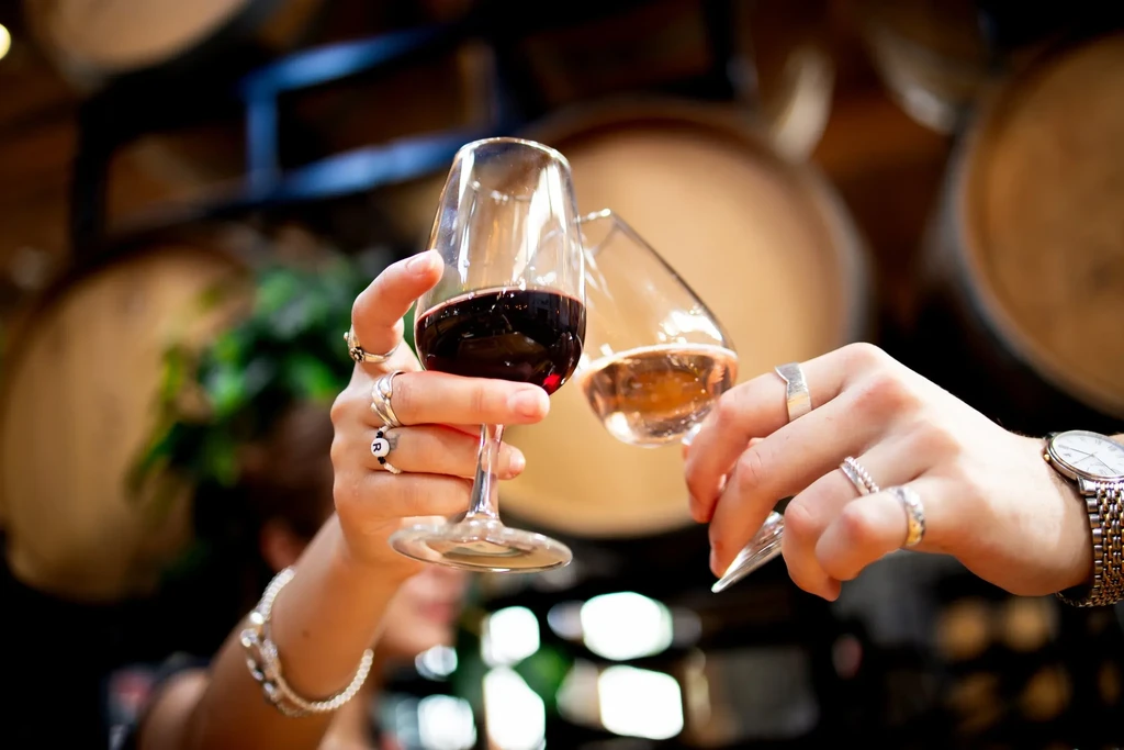 Hands toasting with red and white wine glasses, winery background.