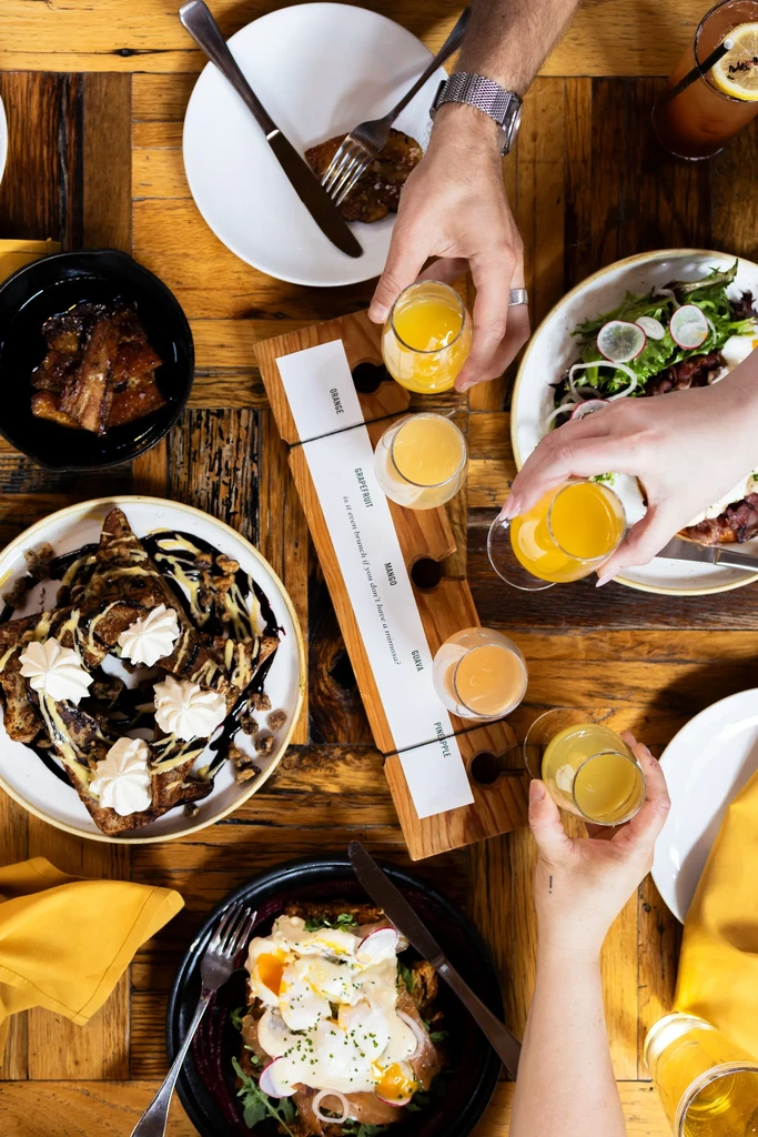 Vibrant brunch spread on rustic table with mimosa flight, hands reaching.