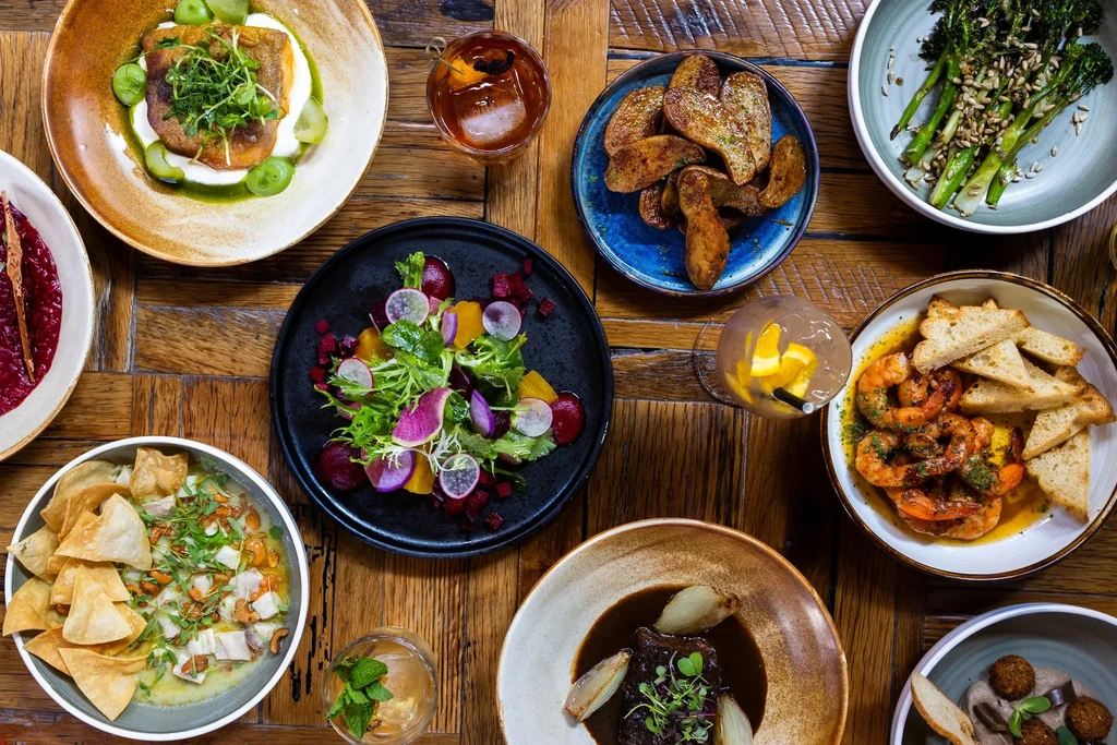 Flat lay of diverse, professionally styled food and cocktails on wood.