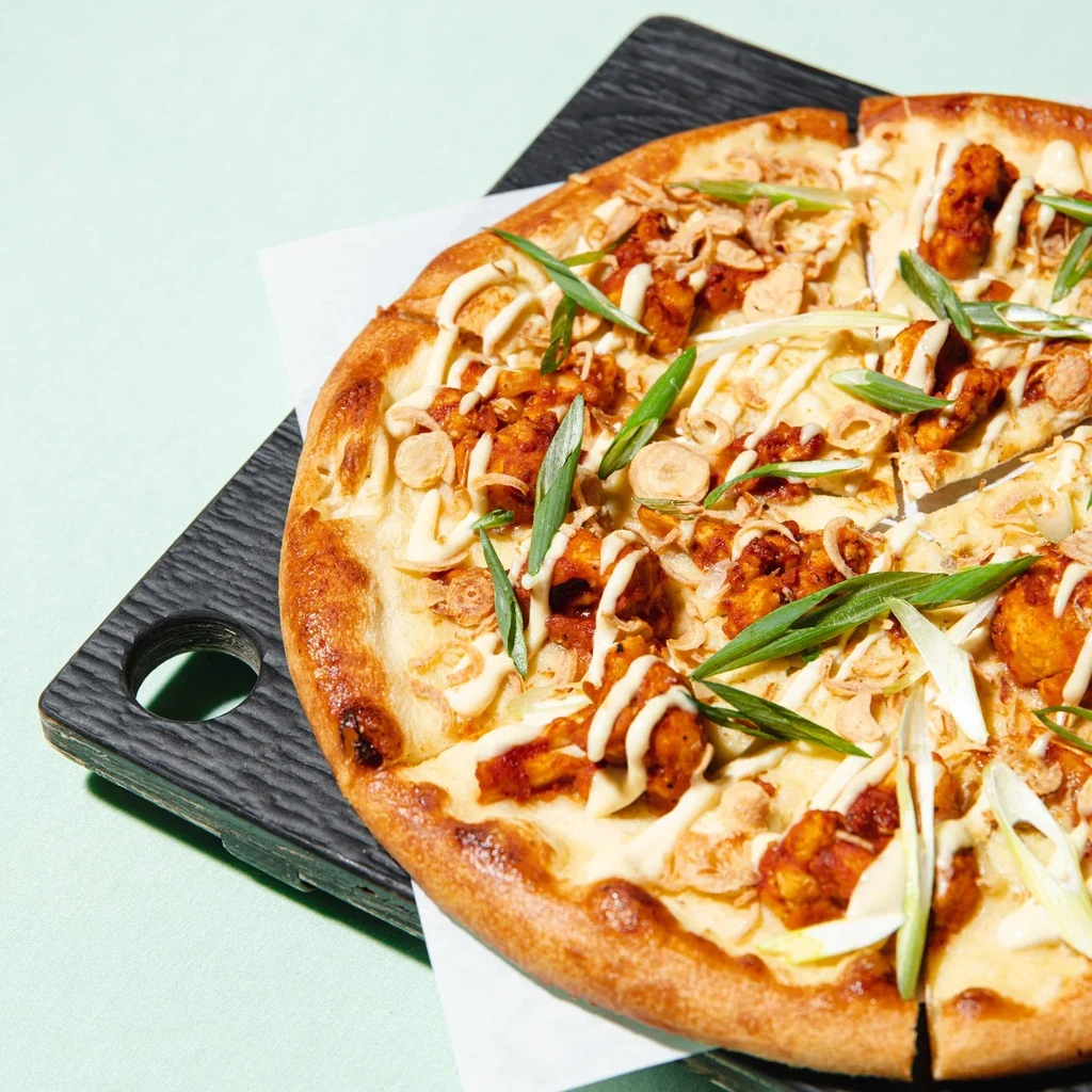 Close-up fried chicken pizza with creamy sauce and scallions on a wooden board.
