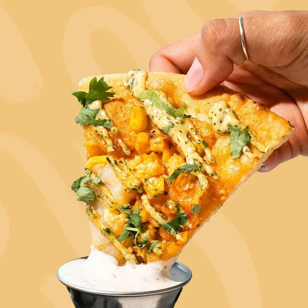 Hand dips shrimp and corn pizza with cilantro into creamy sauce.