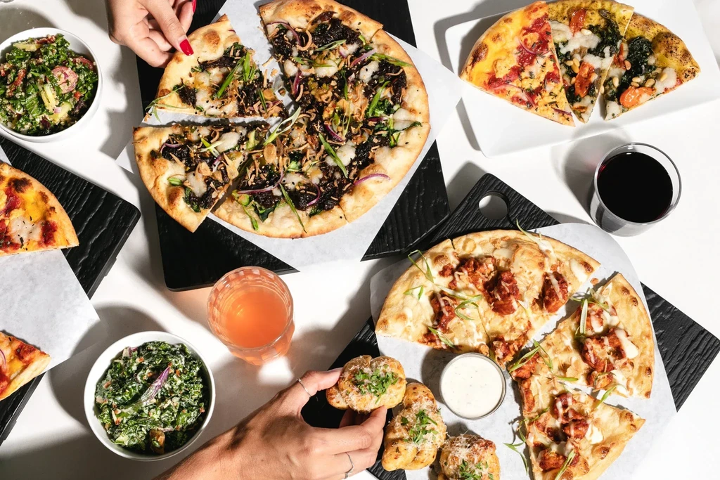 Overhead view of pizzas, salads, drinks, and hands picking food.