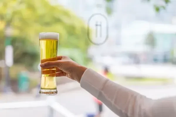 Hand holds glass of golden beer with frothy head, blurred outdoor background.