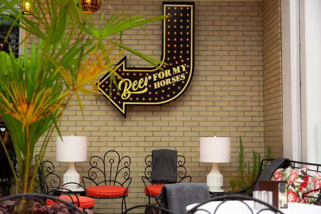 Marquee sign "Beer FOR MY HORSES" over a brick wall patio with furniture.