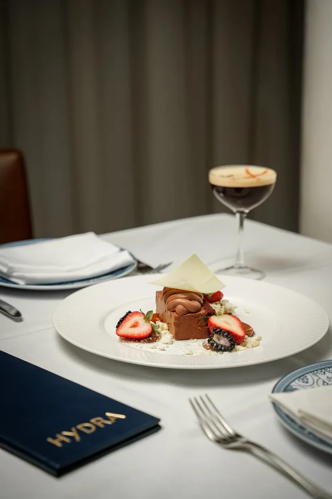 Plated chocolate dessert, dark cocktail, and 'HYDRA' menu on restaurant table.
