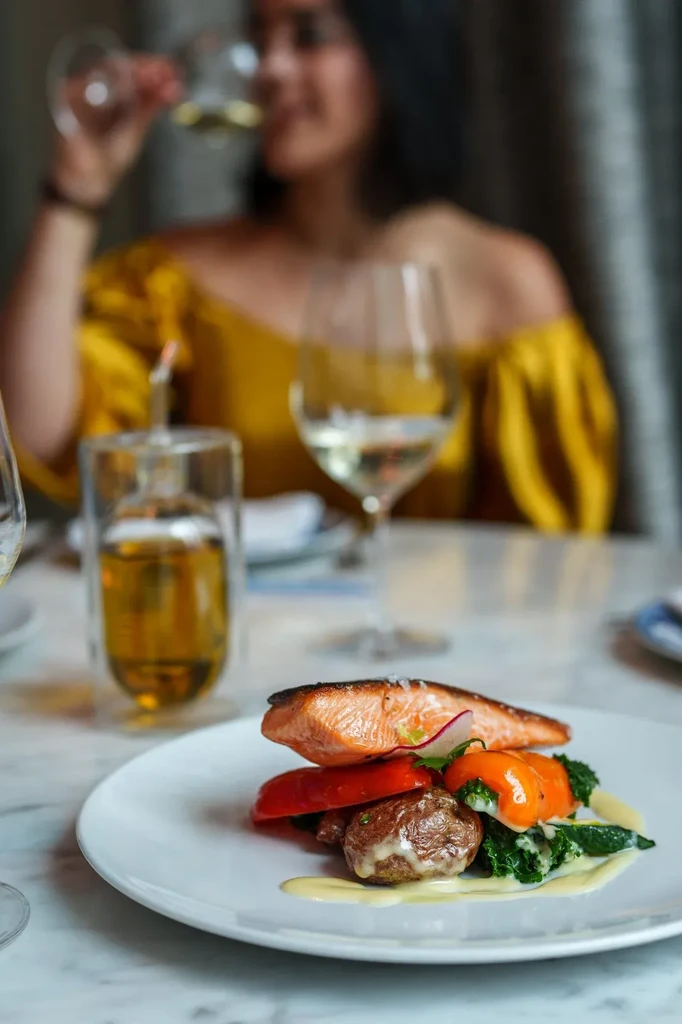 Plated seared salmon, vegetables, wine glasses, blurred person in restaurant.