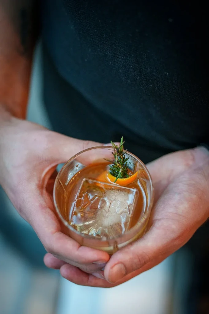 Amber cocktail with orange peel, rosemary, and "HYDRA" embossed ice.