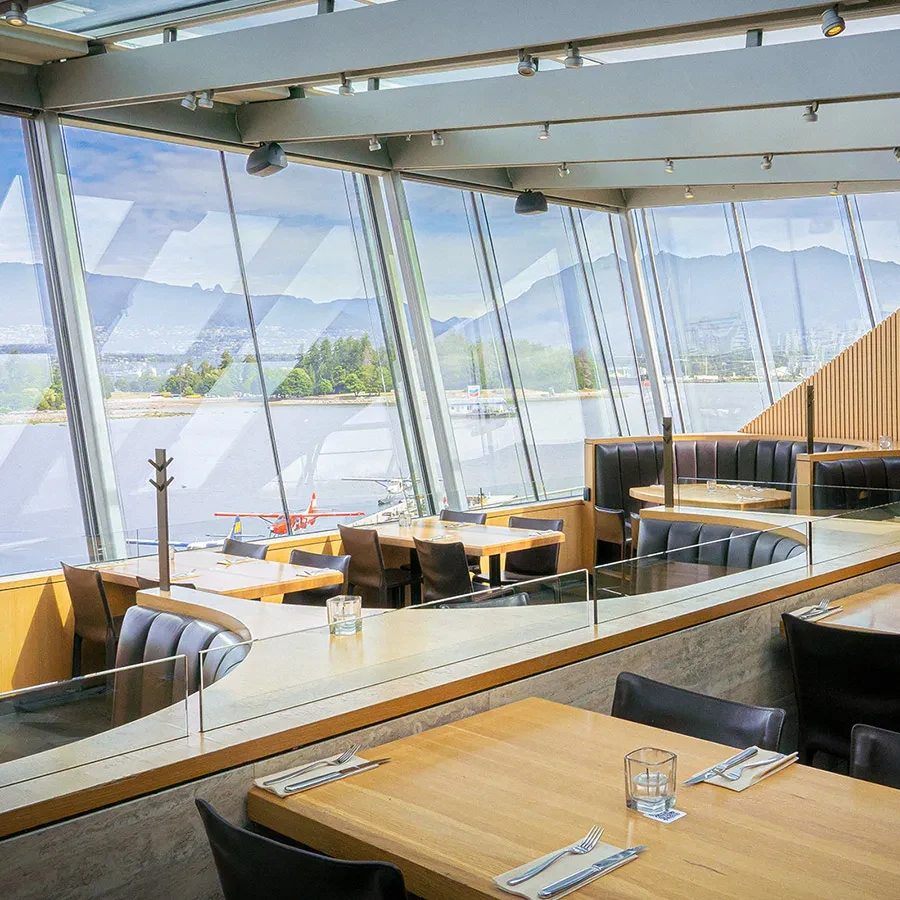 Empty, modern restaurant interior overlooking water, mountains, and cityscape.