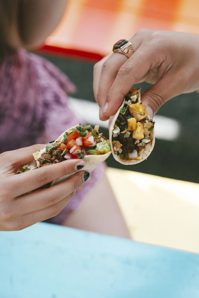 Two hands hold two distinct tacos, one with visible pineapple.