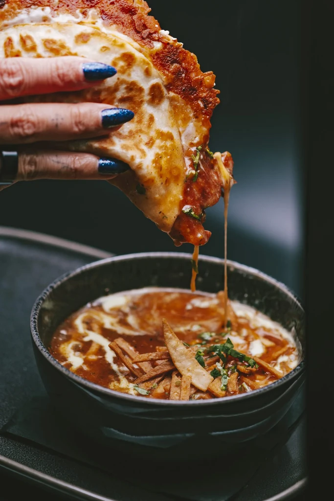 Hand holds cheesy taco dripping into creamy, herbed soup.