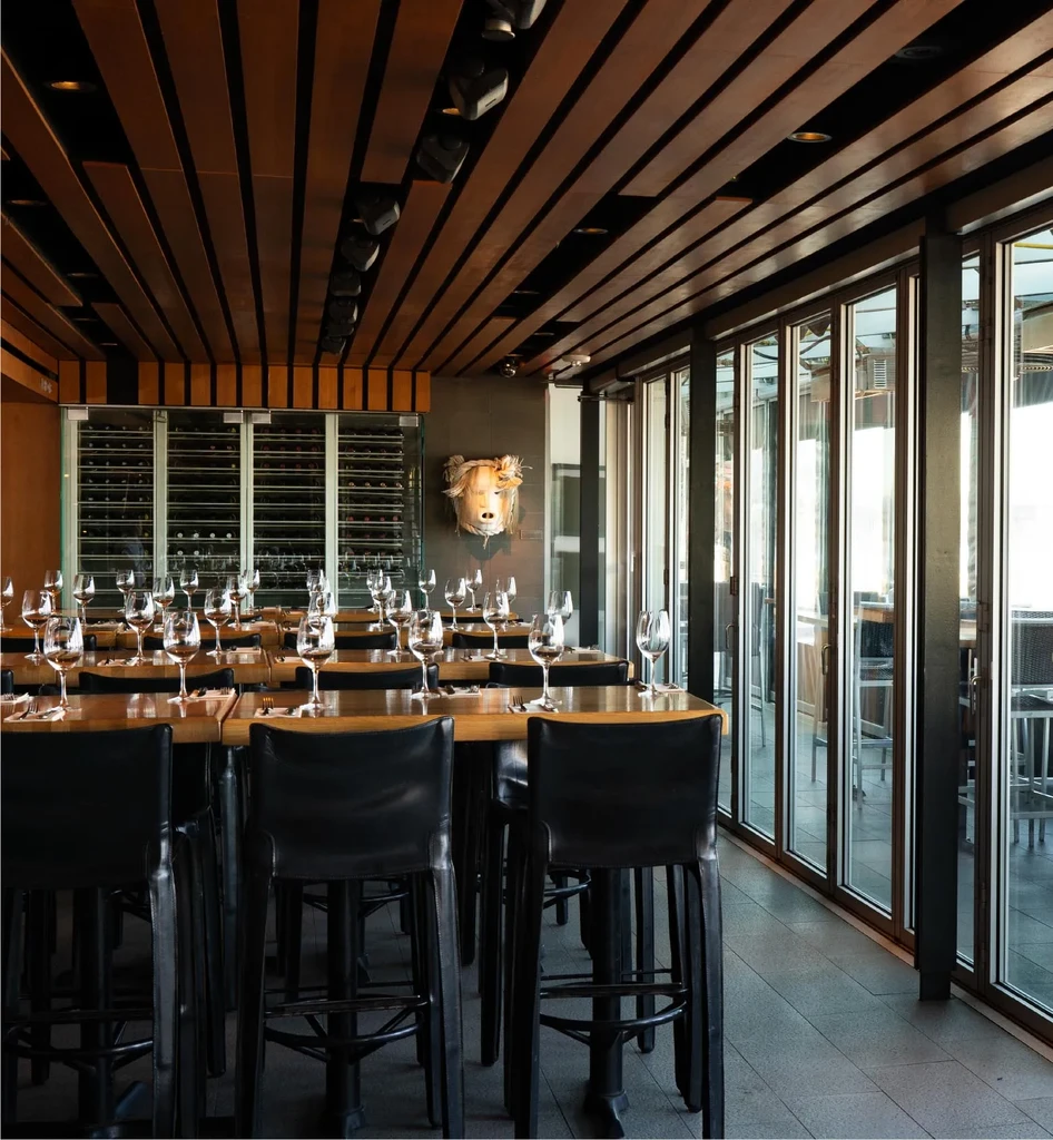 Modern restaurant interior: high-top tables, wine glasses, wood ceiling, wine display.