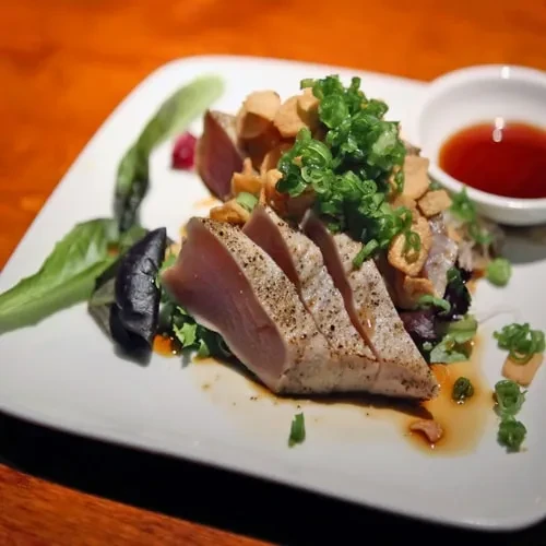 Close-up of seared ahi tuna slices, pink interior, greens, scallions, garlic.
