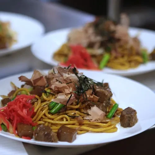 Close-up of yakisoba noodles topped with meat, green vegetables, ginger, and bonito.