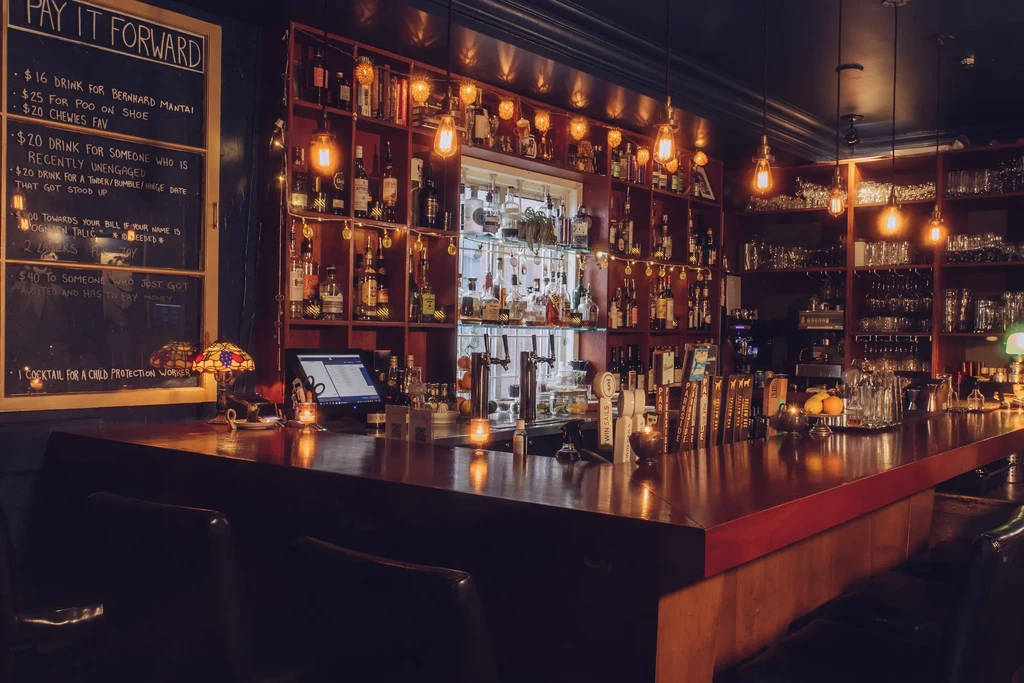 Dimly lit bar interior, wooden counter, shelves of spirits, warm Edison bulbs.