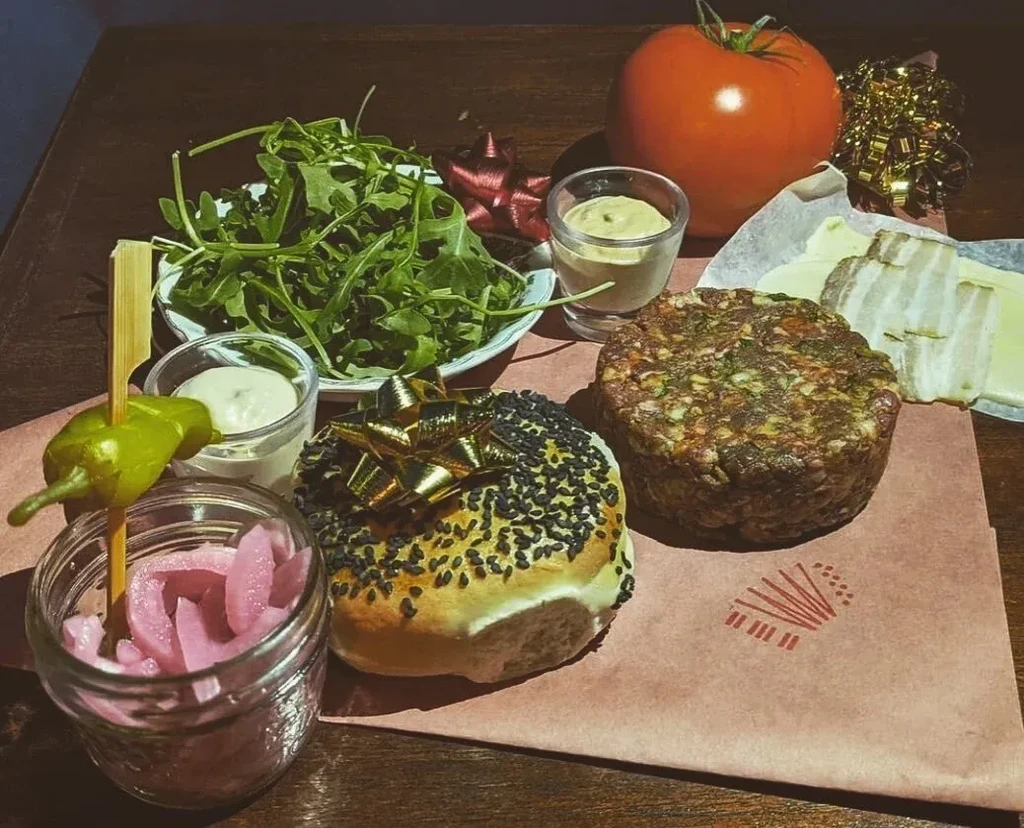 Deconstructed burger platter with sesame bun, thick patty, pickles, and tomato.