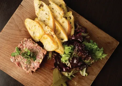 Meat tartare, baguette, mixed greens, and gherkins on a wooden board.